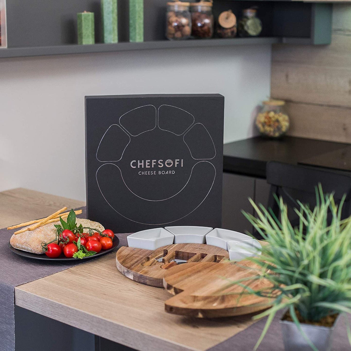 Juodas sūrių padėklas su baltomis lėkštelėmis, medinėmis detalėmis ir šviežiais ingredientais.